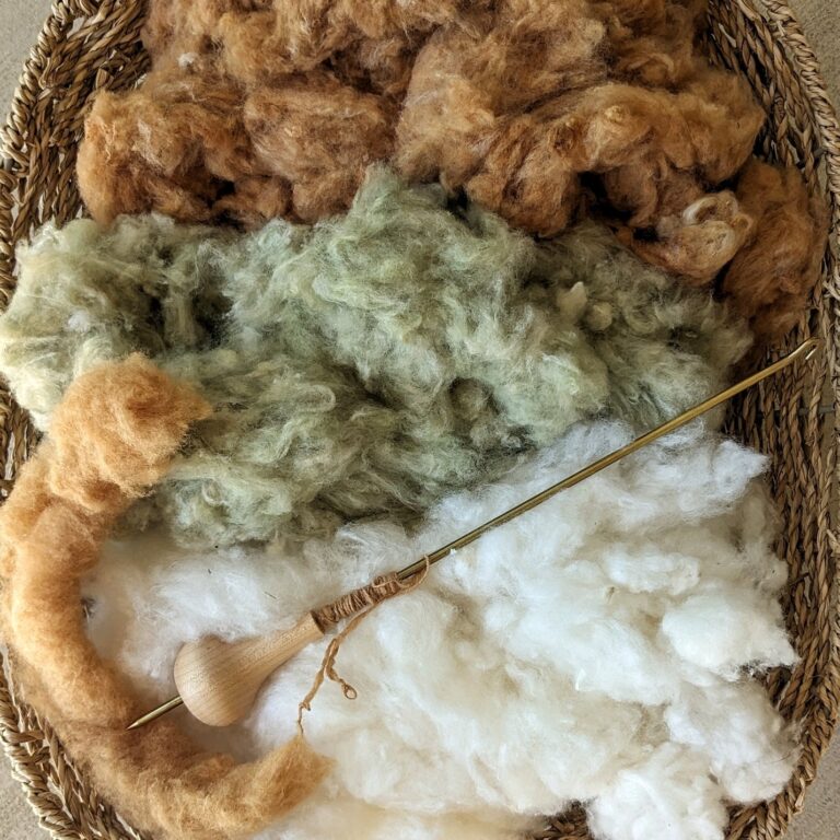 A basket full of three colors of ginned cotton: brown, green, and white. A tahkli rests on top.