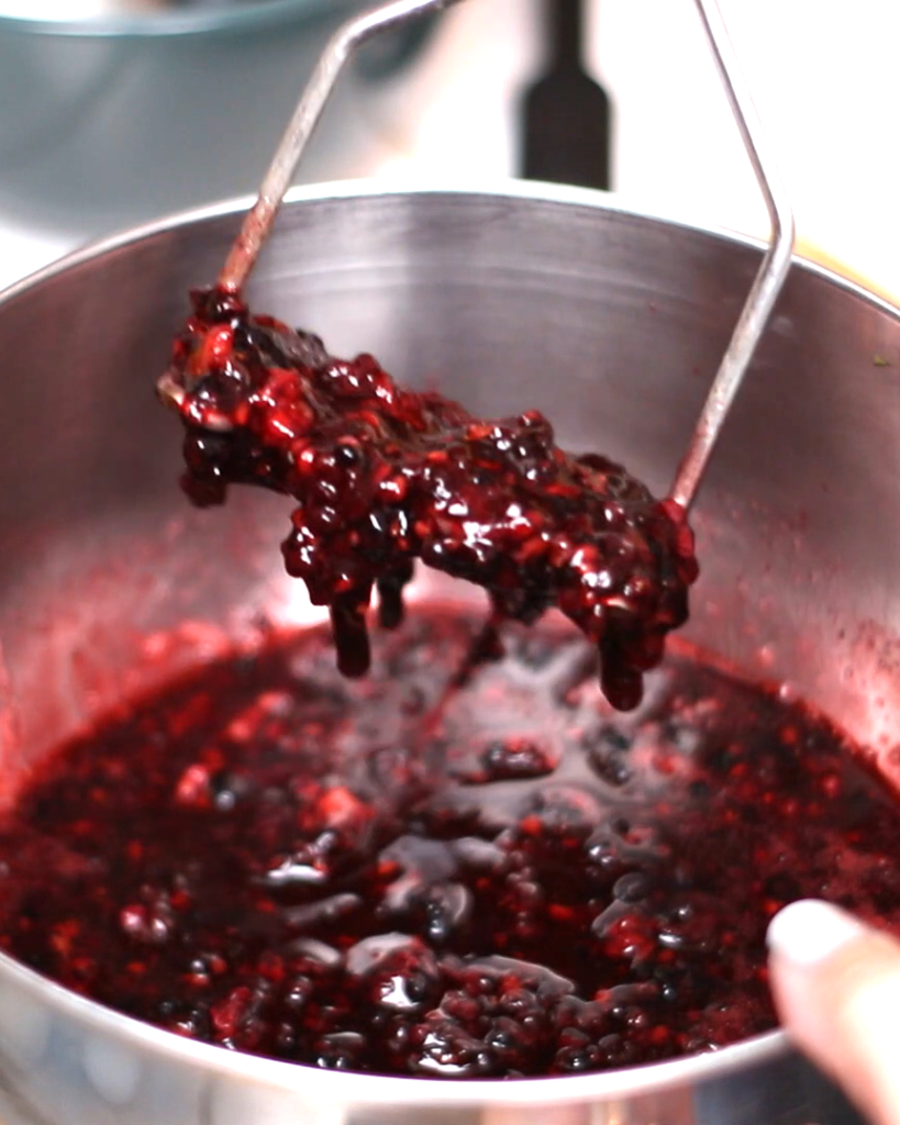 a potato masher crushing a bowl of blackberries and sugar.