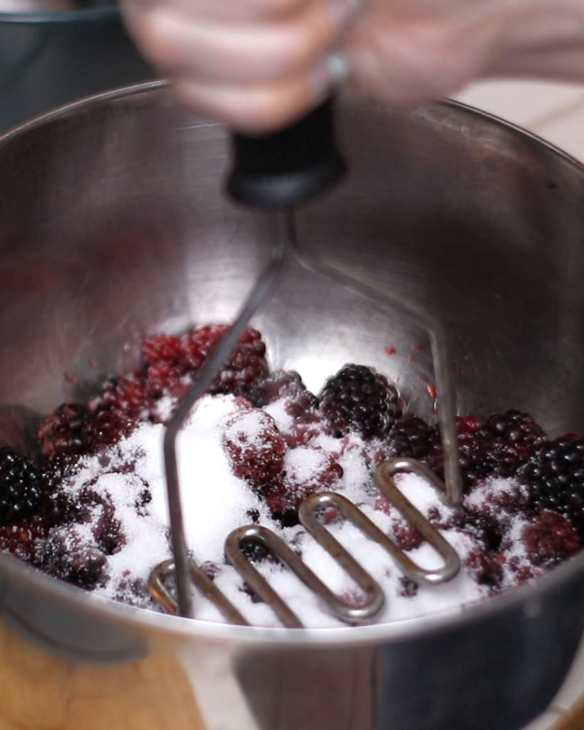a potato masher crushing a bowl of blackberries and sugar.