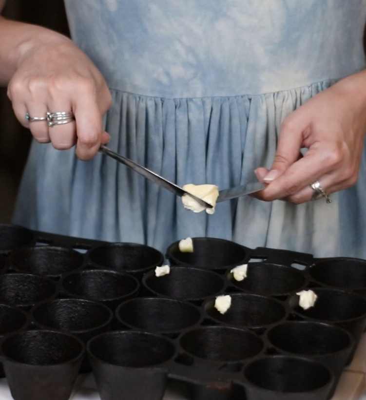 a woman putting a dab of butter into each cup of a cast iron muffin pan