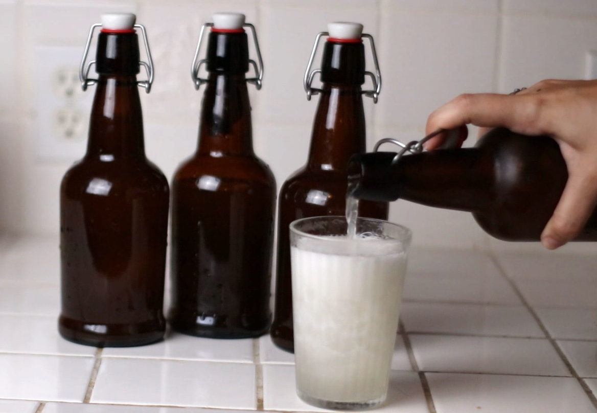 a countertop with three brown glass bottles. a hand is pouring a fourth bottle into a glass of ice. the liquid being poured is ginger beer.