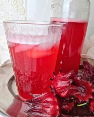 a glass of roselle hibiscus soda next to a glass bottle of the same soda. roselle hibiscus calyxes are next to the glass and bottle.